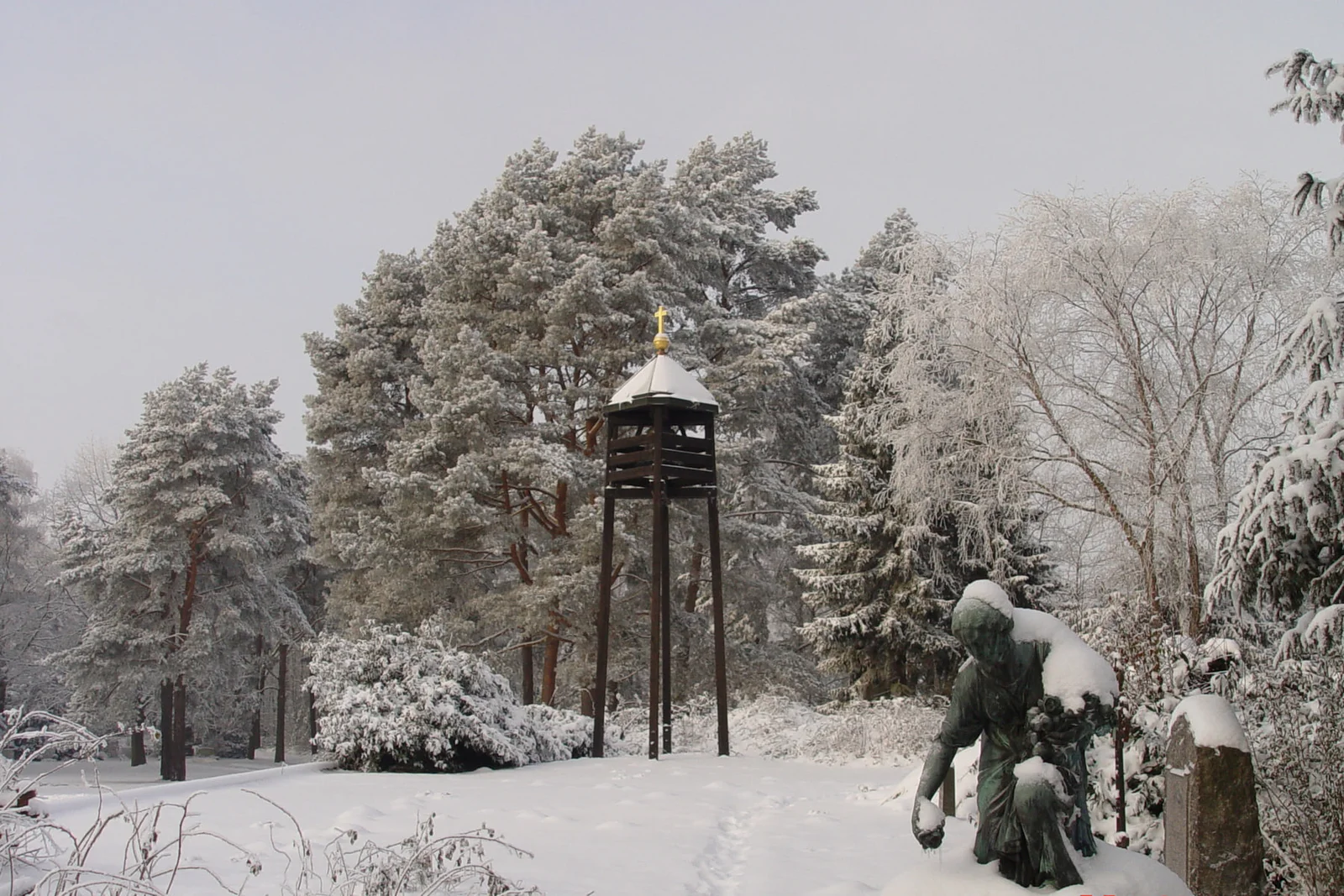 Verschneite Gräber und freistehender dunkler Glockenturm zwischen winterlichen Bäumen auf Friedhof Bergedorf